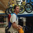 Ein Mann mit buntem Rucksack steht in einem Fahrradladen. Fahrräder hängen über ihm, kleine preisschilder daneben.