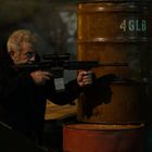 Text "46LB" auf einem Metallfass. Ein älterer Mann zielt mit einem Gewehr, geduckt hinter Fässern, nachts in einer dunklen Umgebung.