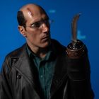 Ein Mann mit Glatze, Narbe auf der Stirn und Brille, trägt Lederjacke, schaut auf Klinge an Stelle einer Hand. Hintergrund blau.