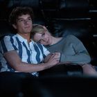 Zwei Personen sitzen eng umschlungen auf dunklen Ledersitzen in einem Kino. Der Mann trägt ein gestreiftes Hemd.