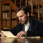 Ein Mann in altmodischer Kleidung liest konzentriert ein Dokument in einer Bibliothek voller Bücher.