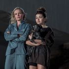 Zwei Frauen schauen ernst, eine trägt einen Hund. Links trägt eine Frau einen blauen Pyjama, rechts hält die andere den Hund.