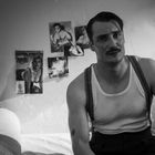 Ein Mann mit Schnurrbart sitzt auf einem Bett in einem schlichten Raum. An der Wand sind Bilder von Muskelmännern und Frauen.