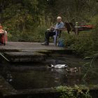 Zwei Personen sitzen auf einer Terrasse am Teich in einem grünen Garten, Enten schwimmen im Wasser.
