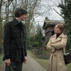 Zwei Personen stehen auf einem Friedhof, umgeben von Bäumen und Grabmälern. Sie tragen Mäntel und führen ein Gespräch.