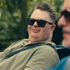 Zwei Männer mit Sonnenbrillen in einem Cabrio, ein Mann lächelt, unscharfer Hintergrund mit Bäumen.