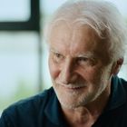 Ich kann diese Person nicht identifizieren. Ein älterer Mann mit weißem Haar und Bart lächelt leicht vor einem Fenster.