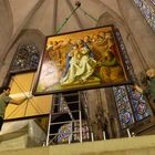 Gemälde von Maria mit Kind, umgeben von Figuren. Es wird in einer Kirche aufgehängt, umrahmt von bunten Glasfenstern.