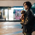 Verschwommenes „PRIMARK“-Logo im Hintergrund, Mann hält einen kleinen, braunen Hund auf einem nächtlichen Platz.