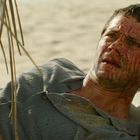 Ein Mann liegt erschöpft und blutig an einem Strand, umgeben von Palmenblättern.