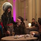 Eine Frau mit großem Fellhut steht lächelnd neben einem sitzenden Mann in einem eleganten, beleuchteten Restaurant.