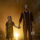 Ein Mann und ein Mädchen stehen in einer nebligen Straße. Sie halten sich an den Händen. Helles Licht im Hintergrund.