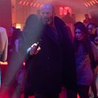 Leuchtendes "ND" in roter Schrift. Eine Person in einem schwarzen Mantel hält etwas in der Hand, umgeben von tanzenden Menschen.