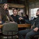 Auf einem Pullover steht "US Marshal". Drei Personen sitzen in einem rustikalen Raum um einen Tisch.