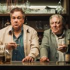 Zwei ältere Männer stehen an einer Bar, halten Gläser. Verschiedene Flaschen und Gläser sind auf der Theke sichtbar.