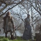 Zwei Männer in mittelalterlicher Gewandung stehen in einem nebligen Wald. Links ein Älterer mit langem Bart und rechts ein Vermummter.
