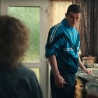 Ein Mann in blauem Trainingsanzug steht in einer Küche, hält einen Teller und schaut zu einer sitzenden Person.