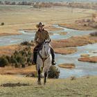 Ein Cowboy reitet auf einem weißen Pferd über einen Hügel, im Hintergrund fließt ein gewundener Fluss durch die Landschaft.
