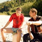 Zwei Männer sitzen auf Steinen im Freien. Der linke trägt ein rotes Sporttrikot, der rechte ein gestreiftes Hemd. Im Hintergrund Bäume.