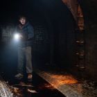 Keine Texte erkennbar. Jemand geht mit einer Taschenlampe durch dunklen, nassen Tunnel aus Ziegeln. Rustikale Metallleiter.