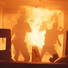 Zwei Personen mit Atemschutzmasken stehen vor einer großen Flamme in einem metallischen Raum voller Rauch.
