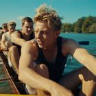 Mehrere Ruderer in einem Boot auf einem See. Der vorderste Ruderer hat blonde Haare und konzentrierten Ausdruck.