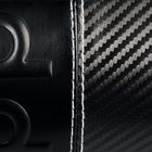 Geprägtes Omega-Symbol links auf schwarzem Leder, rechts Carbonmuster mit weißer Naht dazwischen.