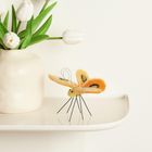 Ein Vase mit weißen Tulpen steht links auf einer glatten, hellen Oberfläche. Rechts davon ein filzener Schmetterling.