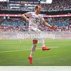 "Vorjahresabbildung" vor einem jubelnden Fußballspieler in weißem Trikot und roten Schuhen, Stadion im Hintergrund.
