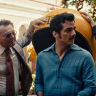 Vier Männer unterhalten sich draußen, ein gelbes Objekt mit der Aufschrift „TELEFONO“ im Hintergrund.