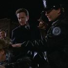 Ein Polizist hält einen Mann auf einem Auto fest; ein anderer beobachtet. Nacht, dramatische Szene.