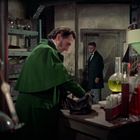 Chemikalien, Glaskolben auf einem Tisch. Zwei Männer in einem Labor; einer im grünen Mantel bei der Arbeit.