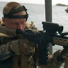 Ein Soldat zielt auf einem Schlauchboot mit einem Gewehr aufs Wasser. Ein weiteres Boot ist im Hintergrund.