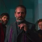 Drei Personen schauen ernst; ein bärtiger Mann in der Mitte mit grauem Anzug, links ein Mann, rechts eine Frau, alle in dunkler Kleidung.