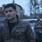 Ein Mann in brauner Lederjacke steht nachdenklich auf der Straße. Im Hintergrund ist eine Frau mit Handy und geöffnetem Auto zu sehen.