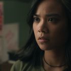 Eine Frau mit ernstem Blick, im Hintergrund unscharfe Poster und ein Schreibtisch.