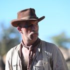 Kein Text. Ein Mann in einem braunen Hut mit Bart und verletztem Gesicht schaut nach rechts, trägt Hemd und Hosenträger.