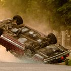 Ein Auto überschlägt sich; Staub wirbelt auf. Links ein Warndreieck, rechts eine weiße Mauer mit Bögen im Hintergrund.