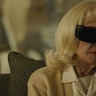 Eine ältere Frau mit blonder Frisur, großen schwarzen Sonnenbrillen und rotem Lippenstift sitzt ernst auf einem Sofa.