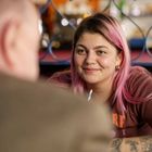 Eine Frau mit rosa Haaren lächelt, tätowierte Arme, trägt ein braunes T-Shirt mit Schriftzug, sitzt in einem Café.