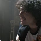 Ein junger Mann mit lockigem Haar lächelt, während er einer Frau mit schwarz-weiß bemaltem Gesicht gegenübersitzt.