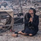 Eine Frau sitzt lächelnd im Schutt mit einem Pelzmantel, umgeben von Trümmern eines zerstörten Gebäudes.