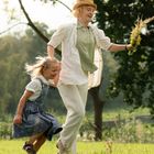 Ein Erwachsener und ein Kind laufen lachend über eine Wiese, der Erwachsene hält einen Blumenstrauß. Im Hintergrund Natur und Bäume.