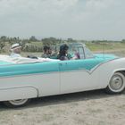 "Sunliner" auf einem weißen und türkisfarbenen Cabrio, das in einer weiten, grünen Landschaft steht. Drei Personen sitzen im Auto.