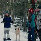 Mehrere Menschen stehen im Schnee. Jeder trägt warme Winterkleidung. Ein Hund ist anwesend. Bäume im Hintergrund.