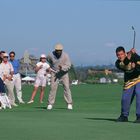 Ein Golfer im schwarzen Trikot schlägt den Ball, umgeben von Zuschauern vor blauem Himmel auf einem Golfplatz.