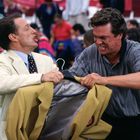 Zwei Männer streiten energisch um ein gelbes Jackett mit einem roten Logo. Menschen in farbenfroher Kleidung im Hintergrund.