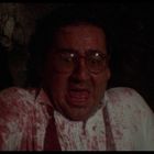 Keine sichtbaren Texte. Ein Mann, erschrocken, mit Brille, blutbeflecktes Hemd, dunkel und beengte Umgebung.