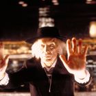 Ein älterer Mann mit weißem Haar und schwarzem Hut streckt beide Hände aus, vor einem unscharfen nächtlichen Stadtpanorama.