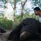 Zwei Personen und ein schlafender Bär im Wald. Eine Person liegt, die andere kniet mit blutigem Tuch.